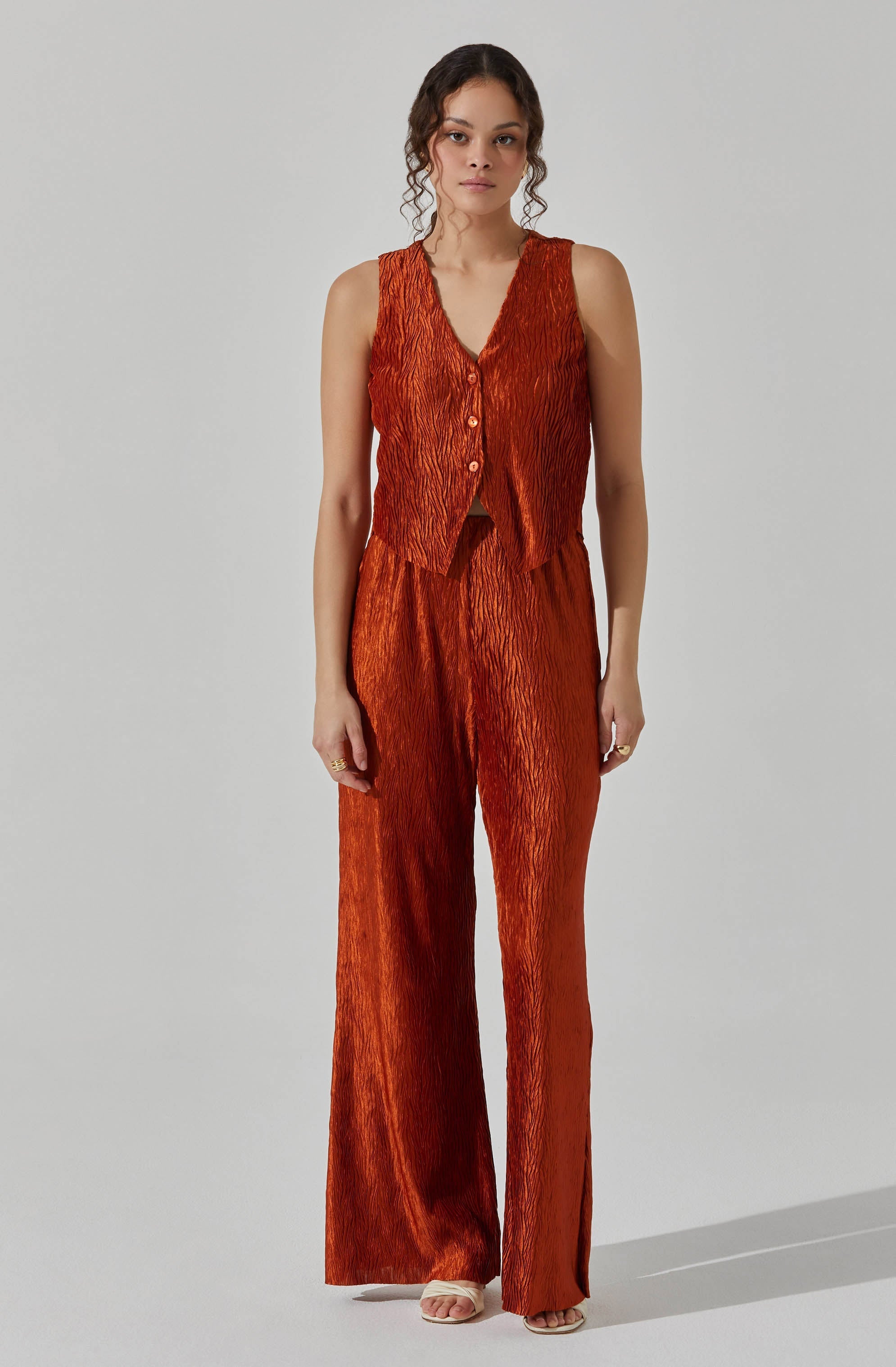 A woman is wearing a matching sleeveless, V-neck vest and wide-leg pants set in a shiny, textured, burnt orange fabric. The vest features small buttons down the front, and the pants are high-waisted with a relaxed fit. She is also wearing open-toe sandals.