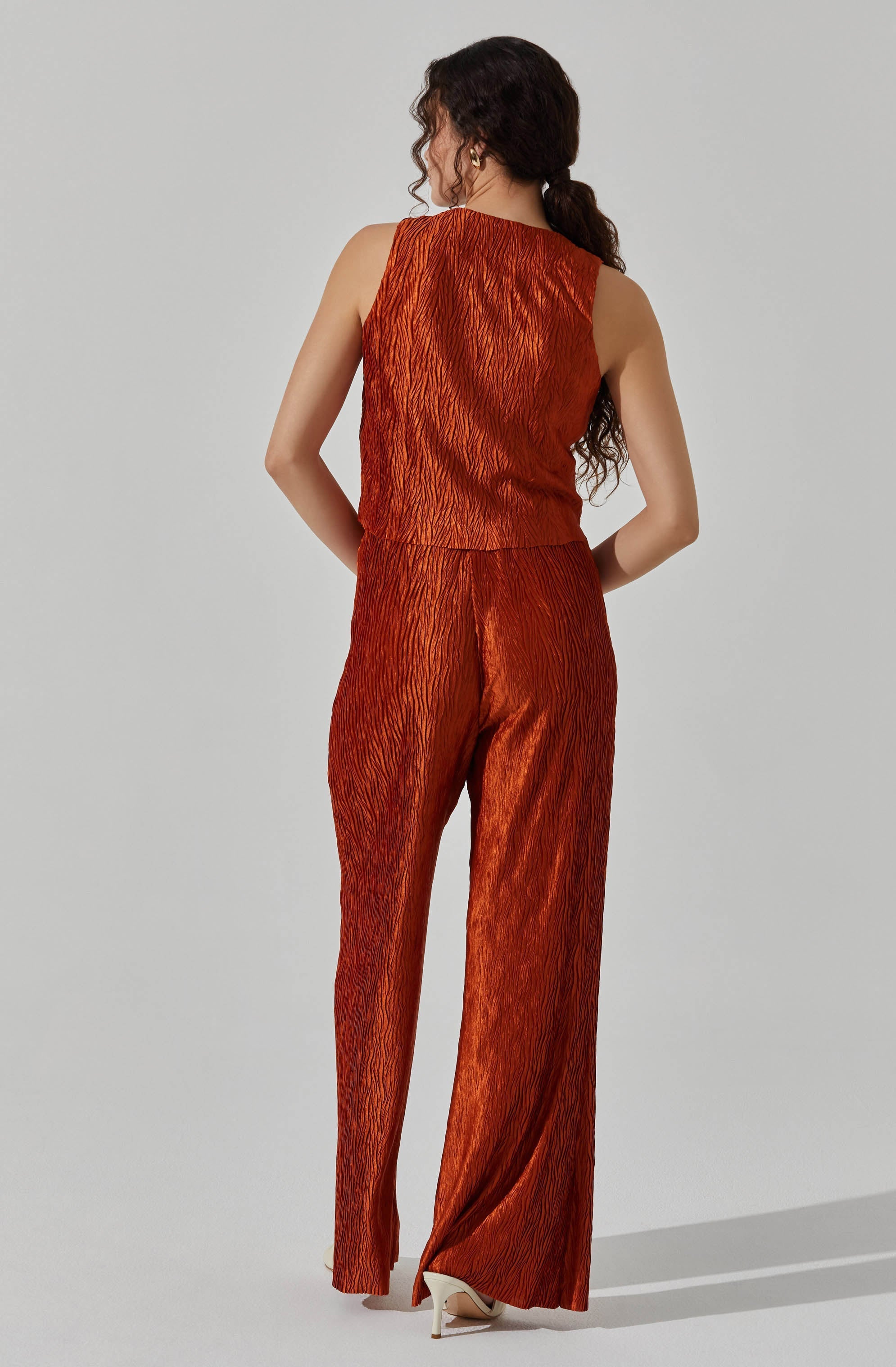 A woman is wearing a sleeveless, rust-colored, textured jumpsuit with wide-leg pants. The fabric has a shiny, crinkled appearance, and she is pairing it with white high-heeled shoes.