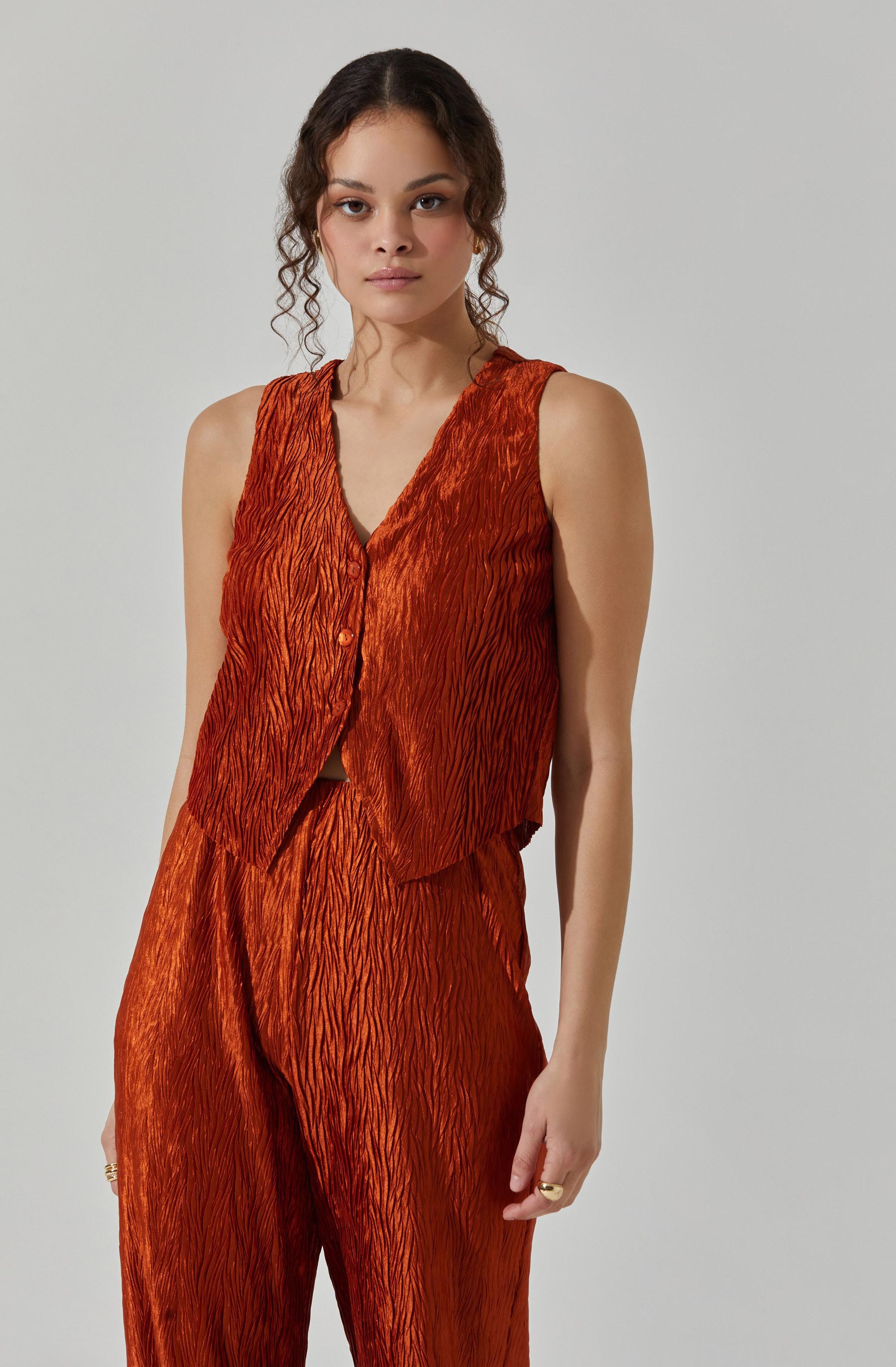 A woman is wearing a sleeveless, burnt orange textured top with a V-neck and button closure in the front. She is also dressed in matching high-waisted pants made from the same textured fabric.
