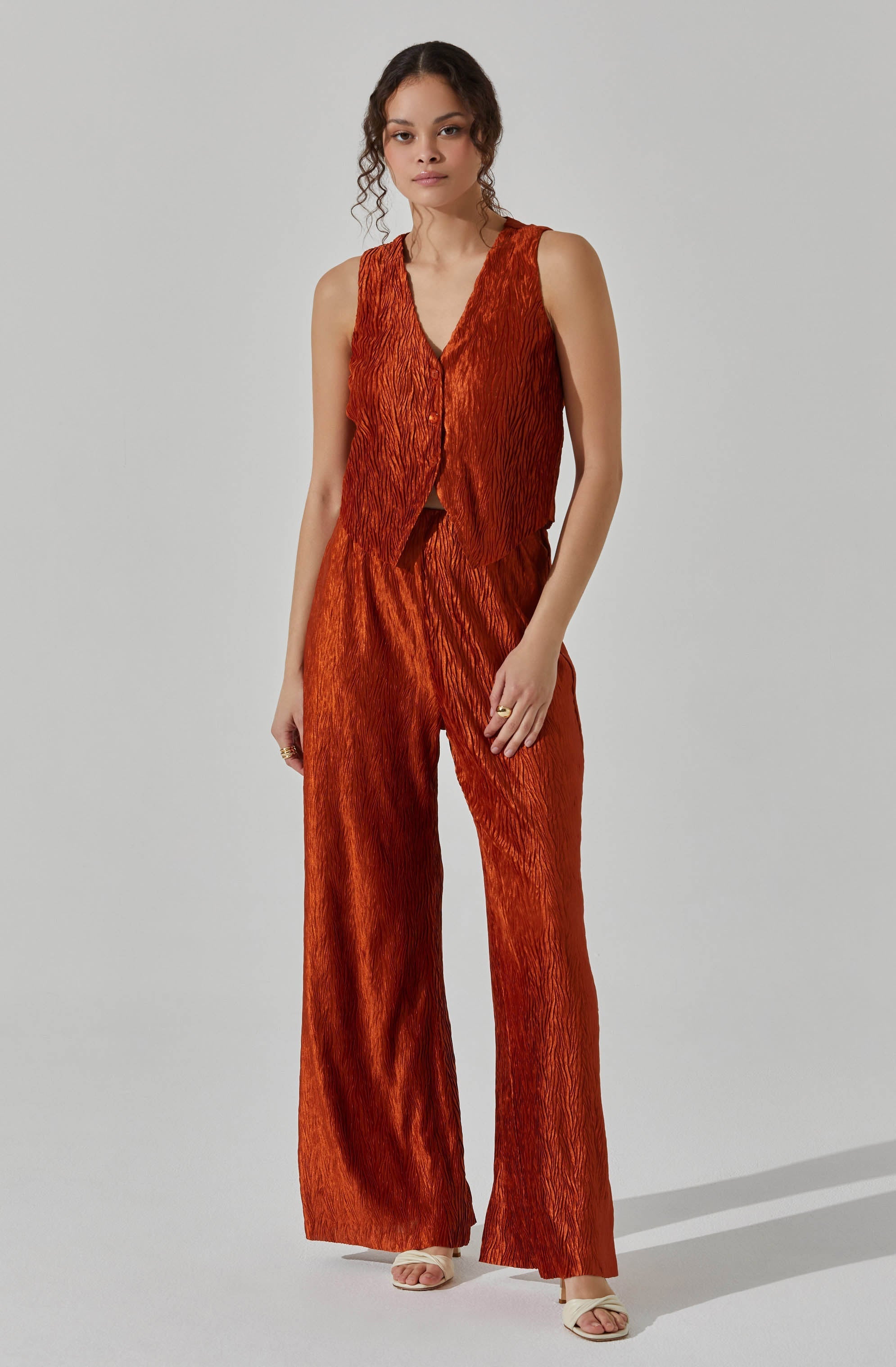 A woman is wearing a sleeveless, V-neck top and matching wide-leg pants, both made of shiny, textured fabric in a rich, burnt orange color. The top has a button-down front, and the pants have a high waist. She is also wearing open-toe, light-colored heels.