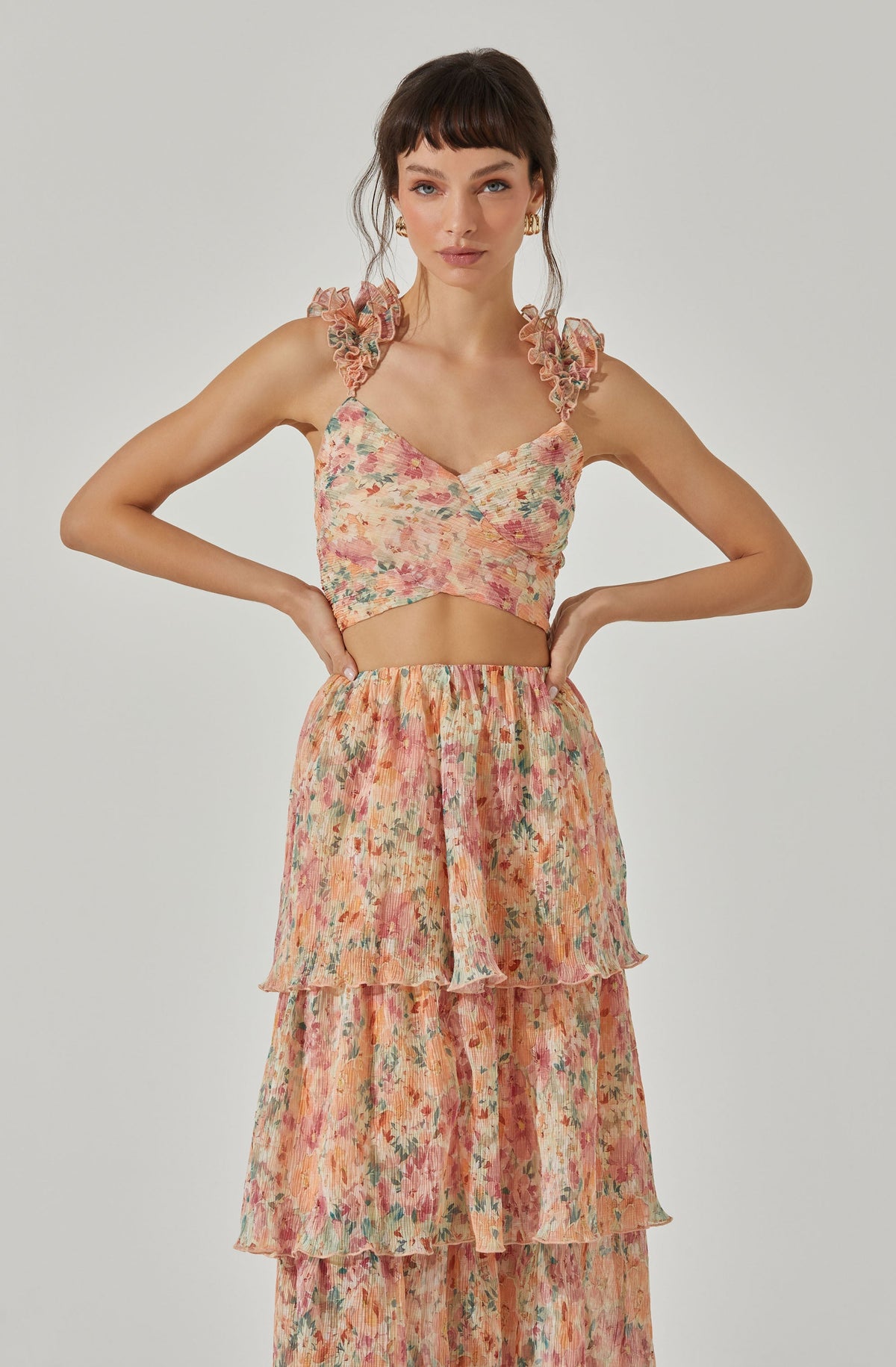 A woman is wearing a sleeveless, floral-patterned crop top with ruffled shoulder straps and a sweetheart neckline. She is also dressed in a high-waisted, tiered, flowy skirt made of lightweight fabric with a matching floral print.