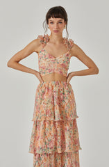 A woman is wearing a sleeveless, floral-patterned crop top with ruffled shoulder straps and a sweetheart neckline. She is also dressed in a high-waisted, tiered, flowy skirt made of lightweight fabric with a matching floral print.