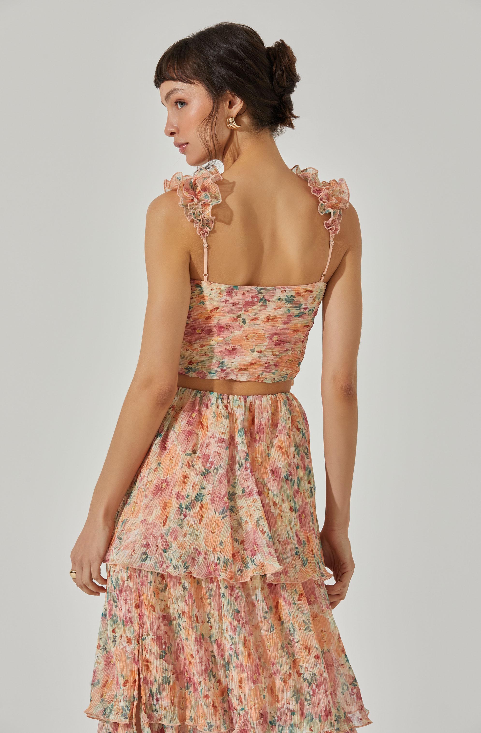 A woman is wearing a two-piece outfit consisting of a sleeveless, fitted crop top with ruffled, sheer shoulder straps, and a high-waisted, tiered skirt. Both the top and skirt feature a colorful, watercolor-style floral pattern in shades of pink, peach, green, and orange.