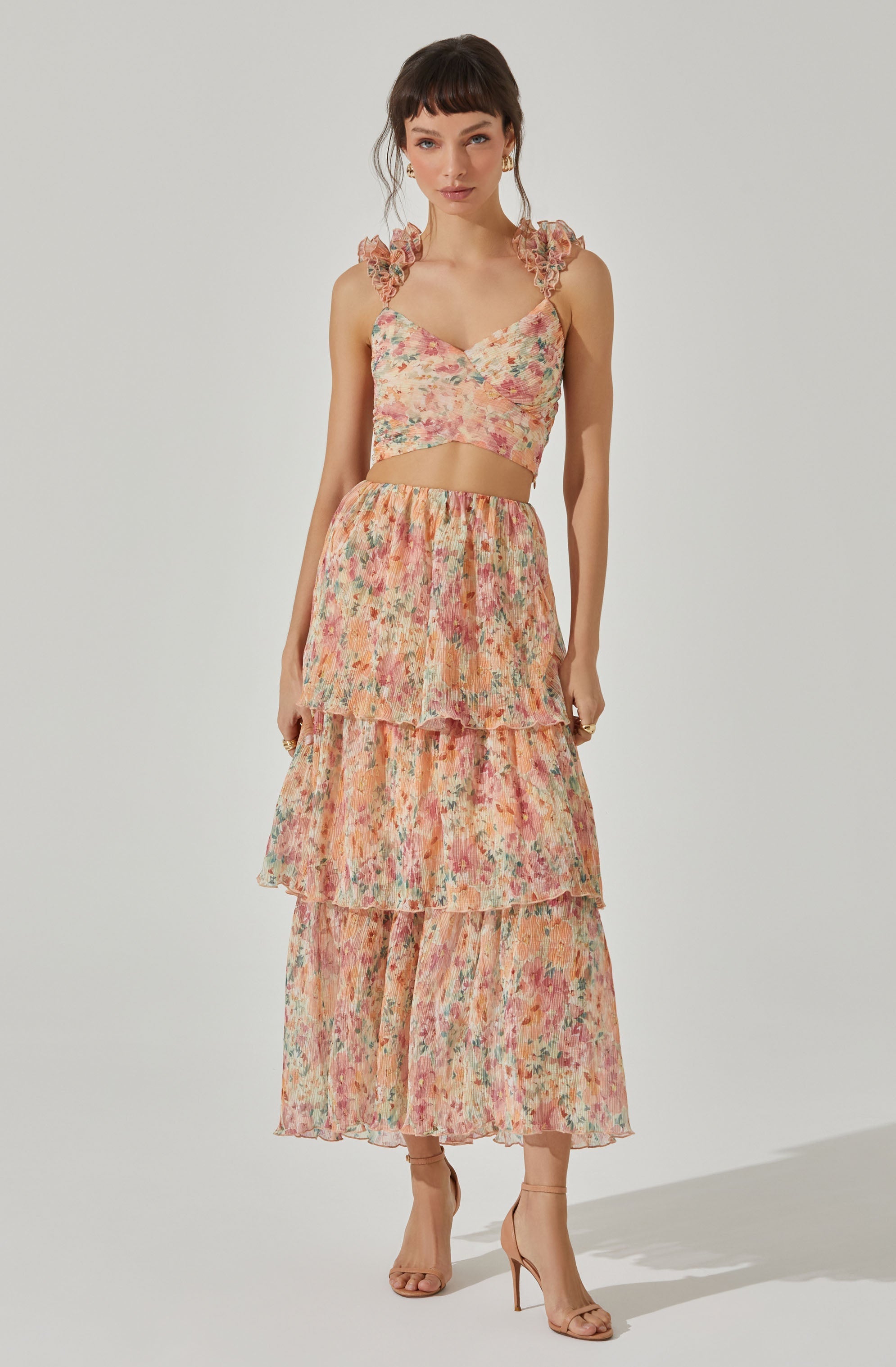 A woman is wearing a matching two-piece outfit, consisting of a sleeveless crop top with ruffled shoulder straps and a long, tiered skirt. The fabric has a colorful, floral pattern with shades of pink, peach, green, and cream. She is also wearing nude high-heeled sandals.