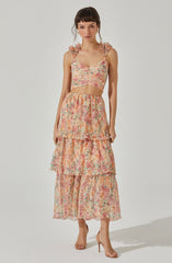 A woman is wearing a matching two-piece outfit, consisting of a sleeveless crop top with ruffled shoulder straps and a long, tiered skirt. The fabric has a colorful, floral pattern with shades of pink, peach, green, and cream. She is also wearing nude high-heeled sandals.