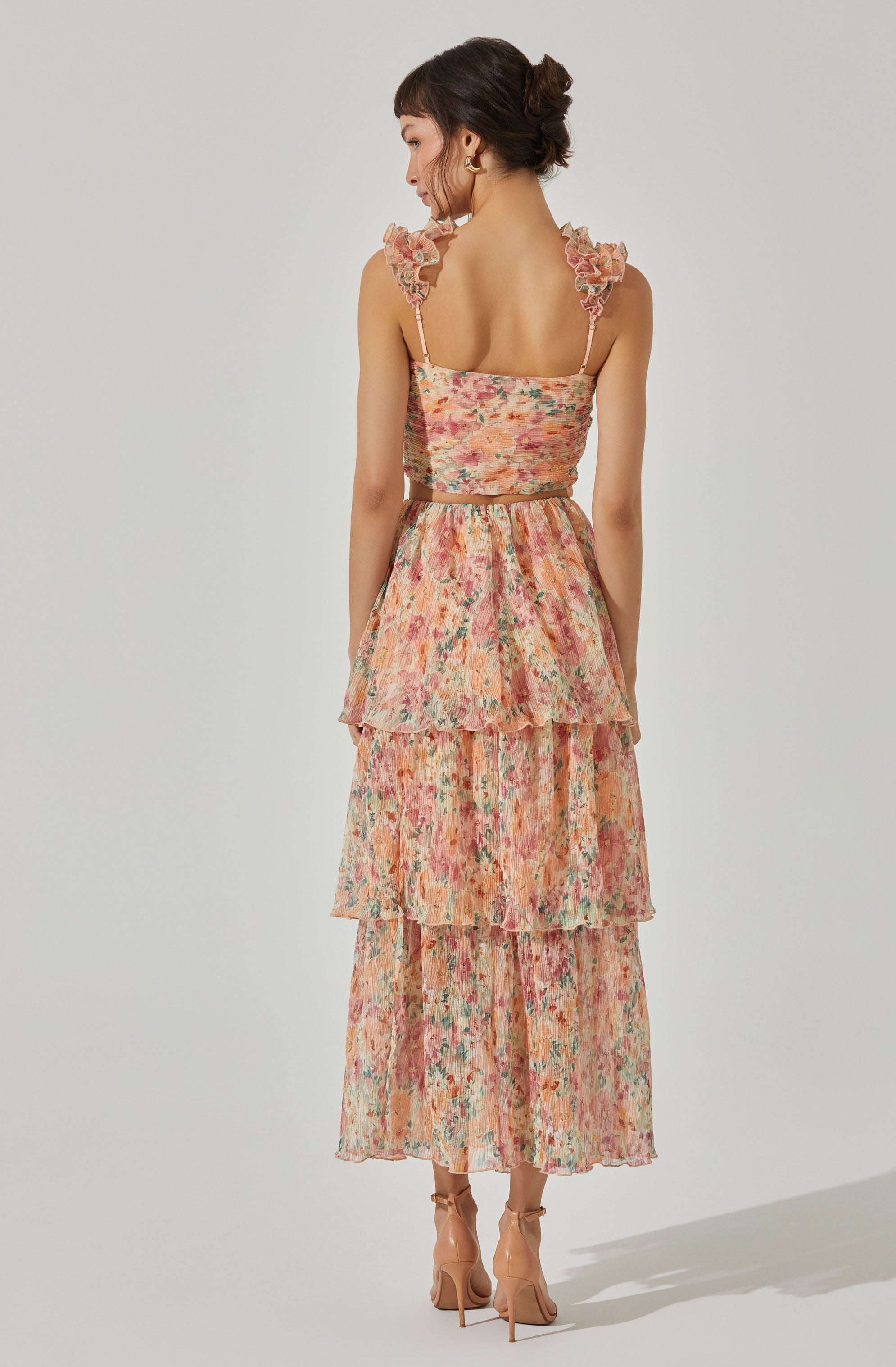 The dress features a sleeveless design with ruffled shoulder straps and a fitted bodice. The fabric has a colorful, watercolor-like floral pattern in shades of pink, orange, green, and beige. The skirt is layered with three tiers of ruffled fabric, creating a flowing, tiered silhouette that reaches mid-calf length. The dress has a soft, feminine appearance with a light, airy texture. The woman is also wearing nude high-heeled sandals.