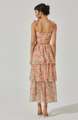 The dress features a sleeveless design with ruffled shoulder straps and a fitted bodice. The fabric has a colorful, watercolor-like floral pattern in shades of pink, orange, green, and beige. The skirt is layered with three tiers of ruffled fabric, creating a flowing, tiered silhouette that reaches mid-calf length. The dress has a soft, feminine appearance with a light, airy texture. The woman is also wearing nude high-heeled sandals.