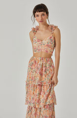 A woman is wearing a two-piece outfit with a sleeveless crop top and a tiered maxi skirt. The crop top has ruffled shoulder straps and a sweetheart neckline, featuring a colorful floral pattern in shades of pink, peach, cream, and green. The matching skirt is high-waisted with an elastic waistband, made of the same floral fabric, and has multiple layered tiers creating a flowing silhouette.