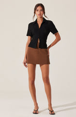 A woman is wearing a short-sleeved black textured button-up shirt with a collar, paired with a high-waisted, A-line brown mini skirt. She is also wearing black open-toe high heels.