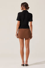 A woman with dark brown hair styled in a low updo is wearing a textured black short-sleeve shirt with a collar. She is also wearing a brown A-line mini skirt with a front seam detail and a side pocket visible on her right side. She completes the outfit with black open-toe heels.