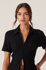 A woman wearing a black textured short-sleeve button-up shirt with a collar. The shirt has a slightly open front, revealing her midriff.