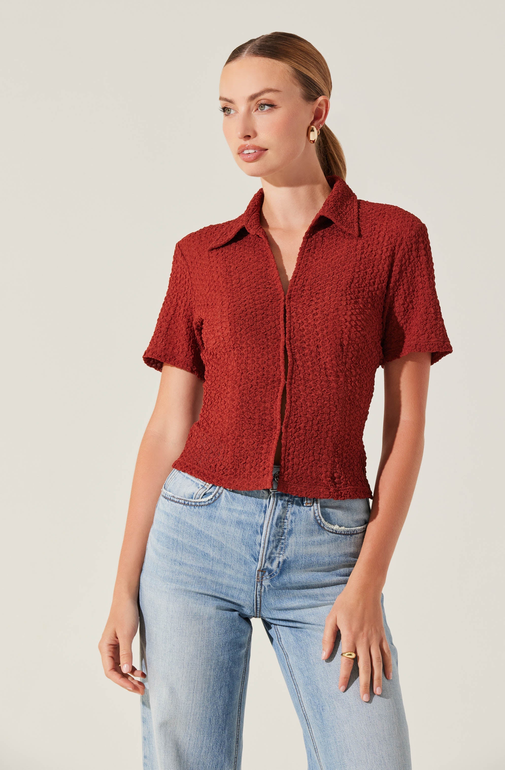 A woman is wearing a short-sleeved, textured red collared shirt with a front zipper, paired with high-waisted light blue jeans.