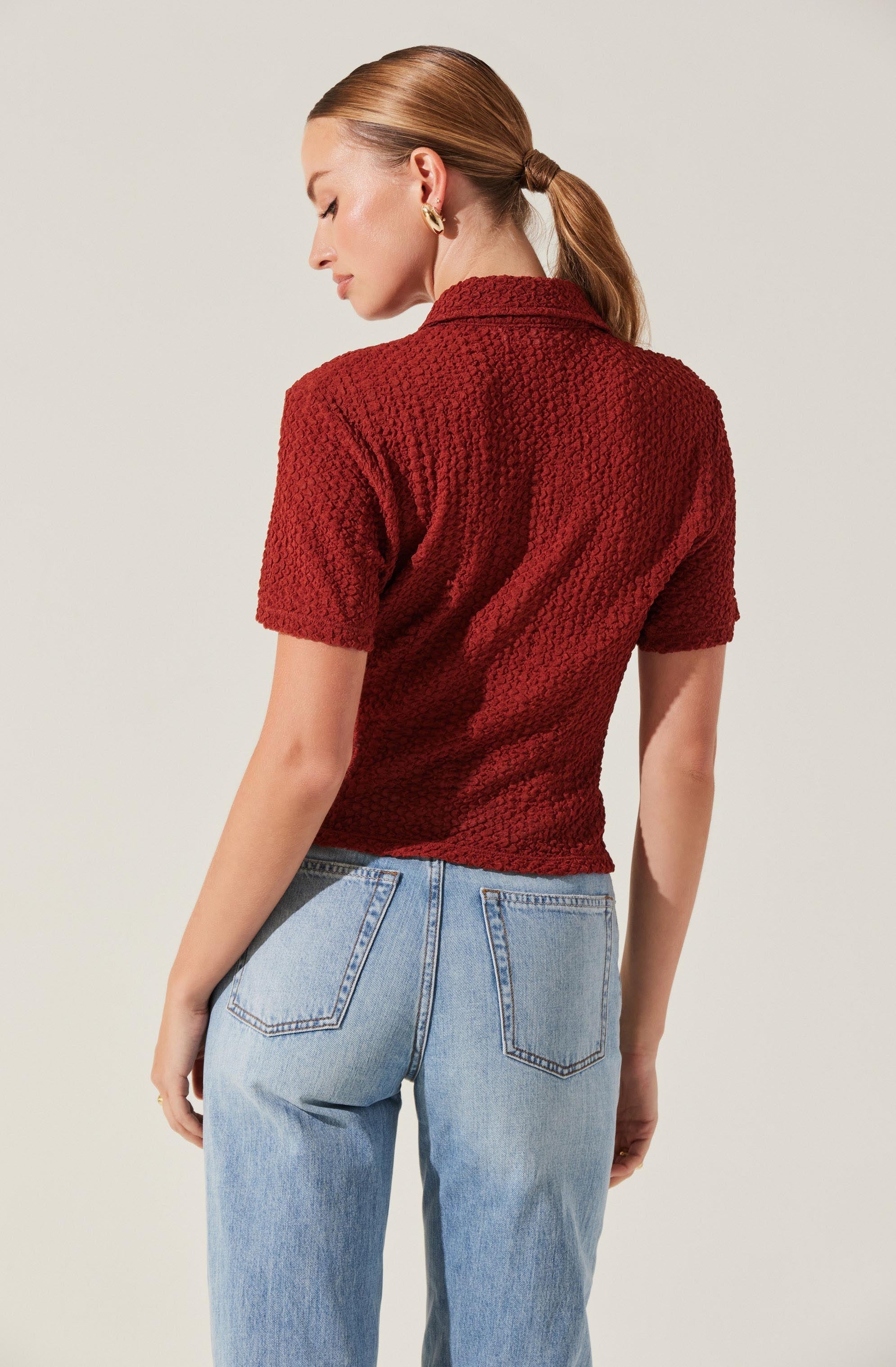 A woman is wearing a textured, short-sleeved, collared red top paired with light blue high-waisted jeans. The top has a slightly loose fit, and the jeans have classic denim styling with visible stitching and back pockets. The woman's hair is styled in a low ponytail, and she is accessorized with gold hoop earrings.
