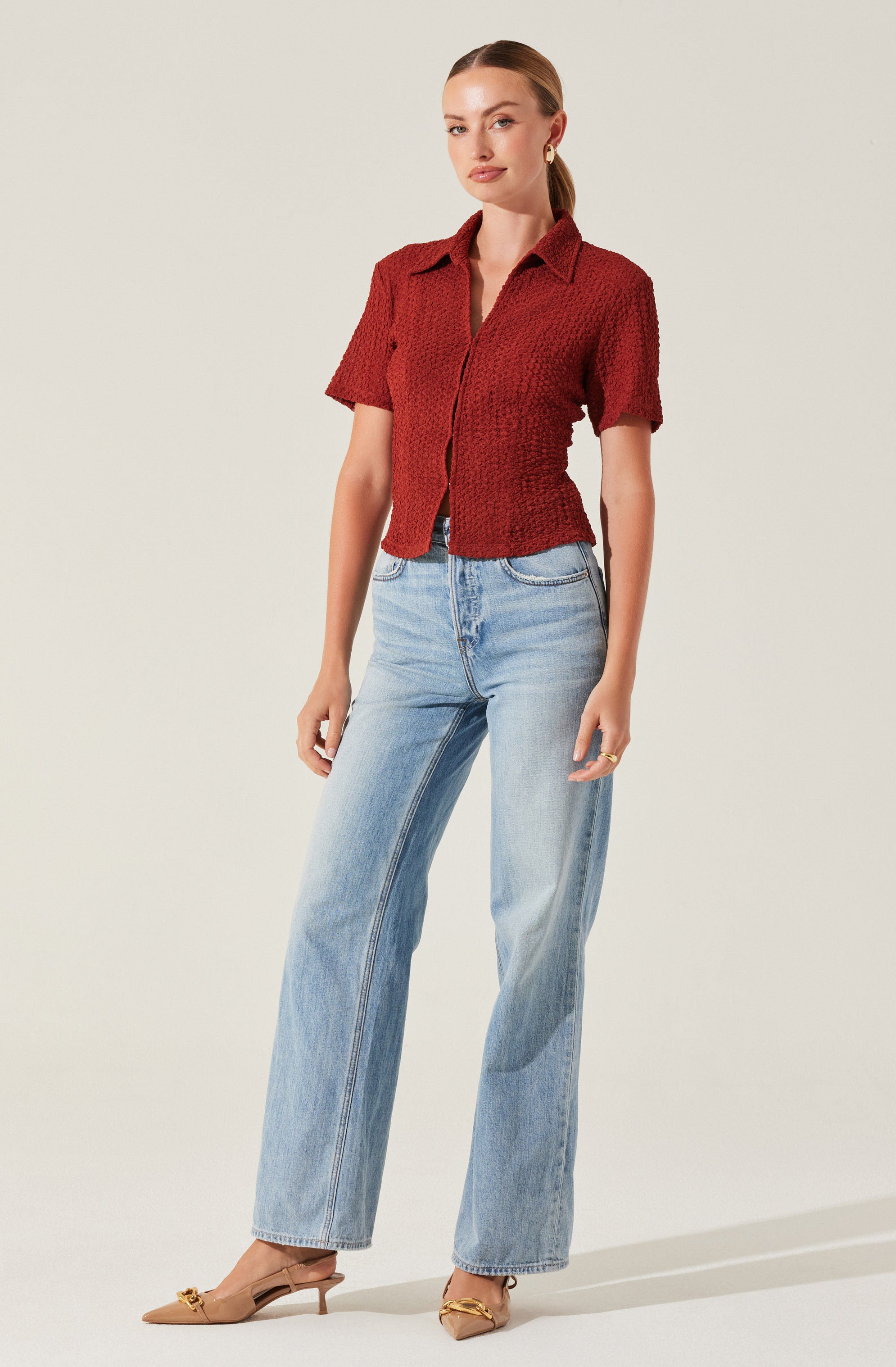 A woman is wearing a textured, short-sleeved, collared red shirt with a front opening. She pairs it with light blue, high-waisted, wide-leg jeans. She is also wearing beige pointed-toe heels with a decorative detail on the front.