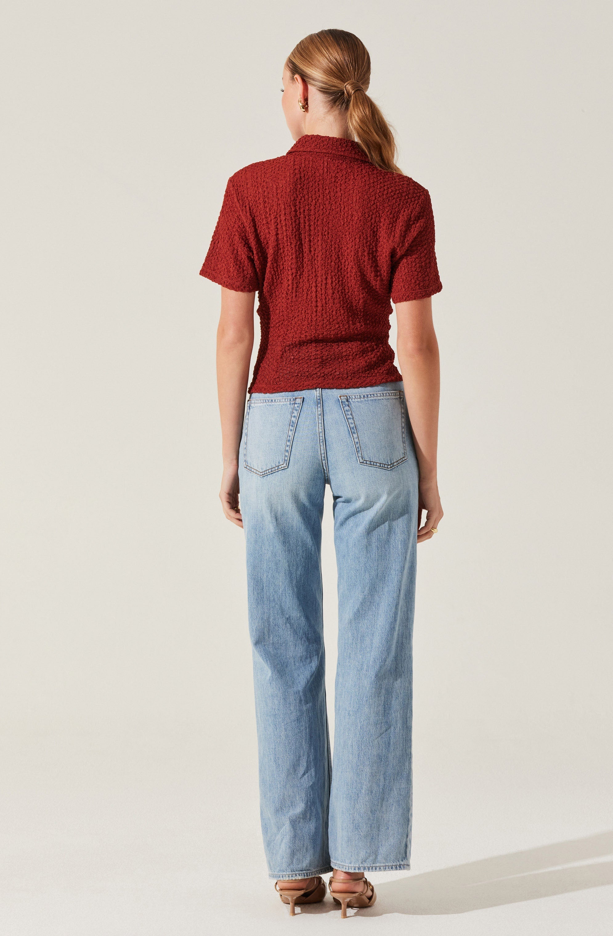 A woman is wearing a short-sleeved, textured red knit top and light blue high-waisted jeans with a relaxed fit. She is also wearing open-toe beige heels.