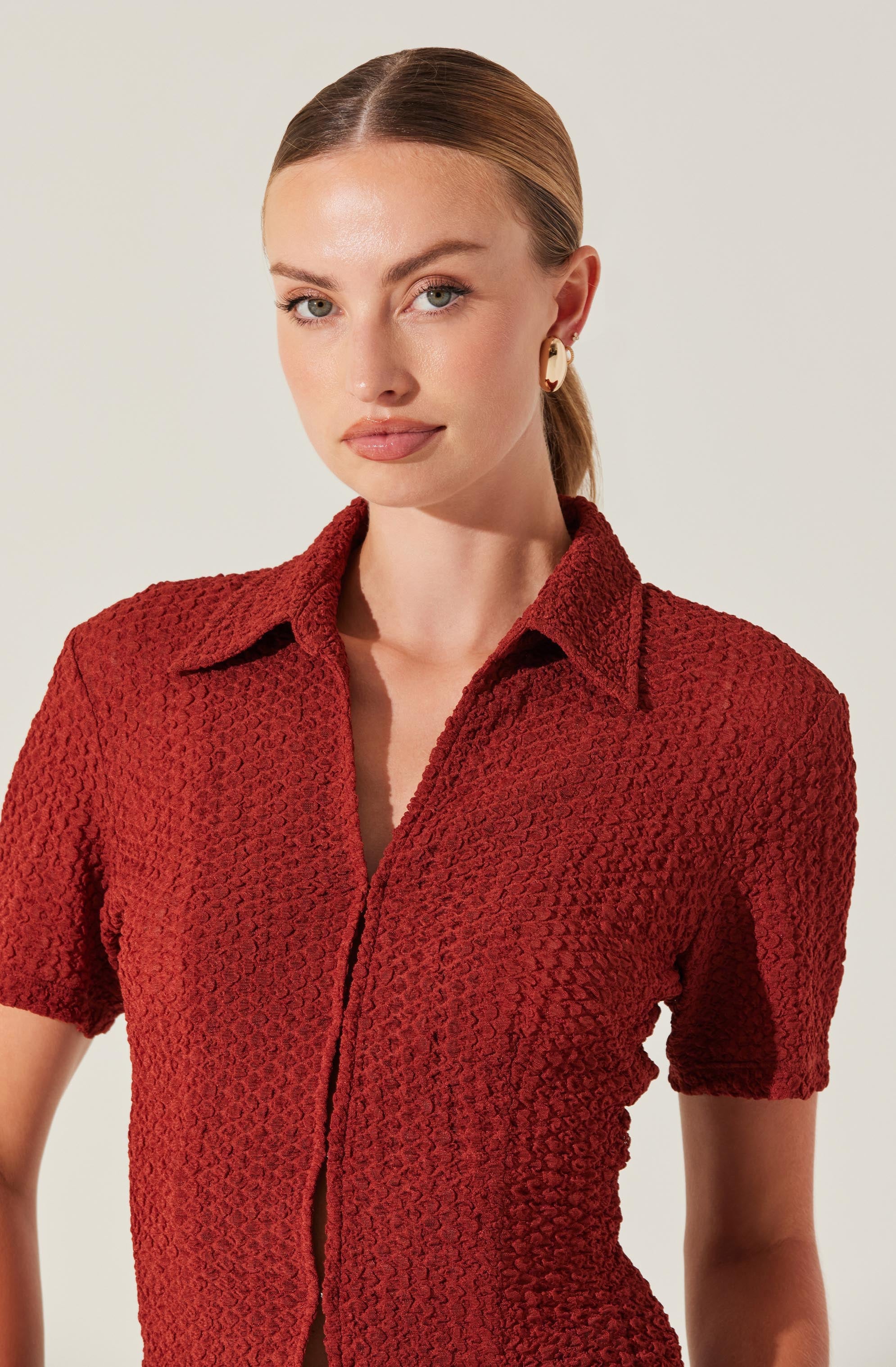 A woman is wearing a textured, short-sleeved, button-up shirt in a deep red color. The shirt features a classic collar and a fitted silhouette. She is accessorized with a pair of large, cream-colored hoop earrings.