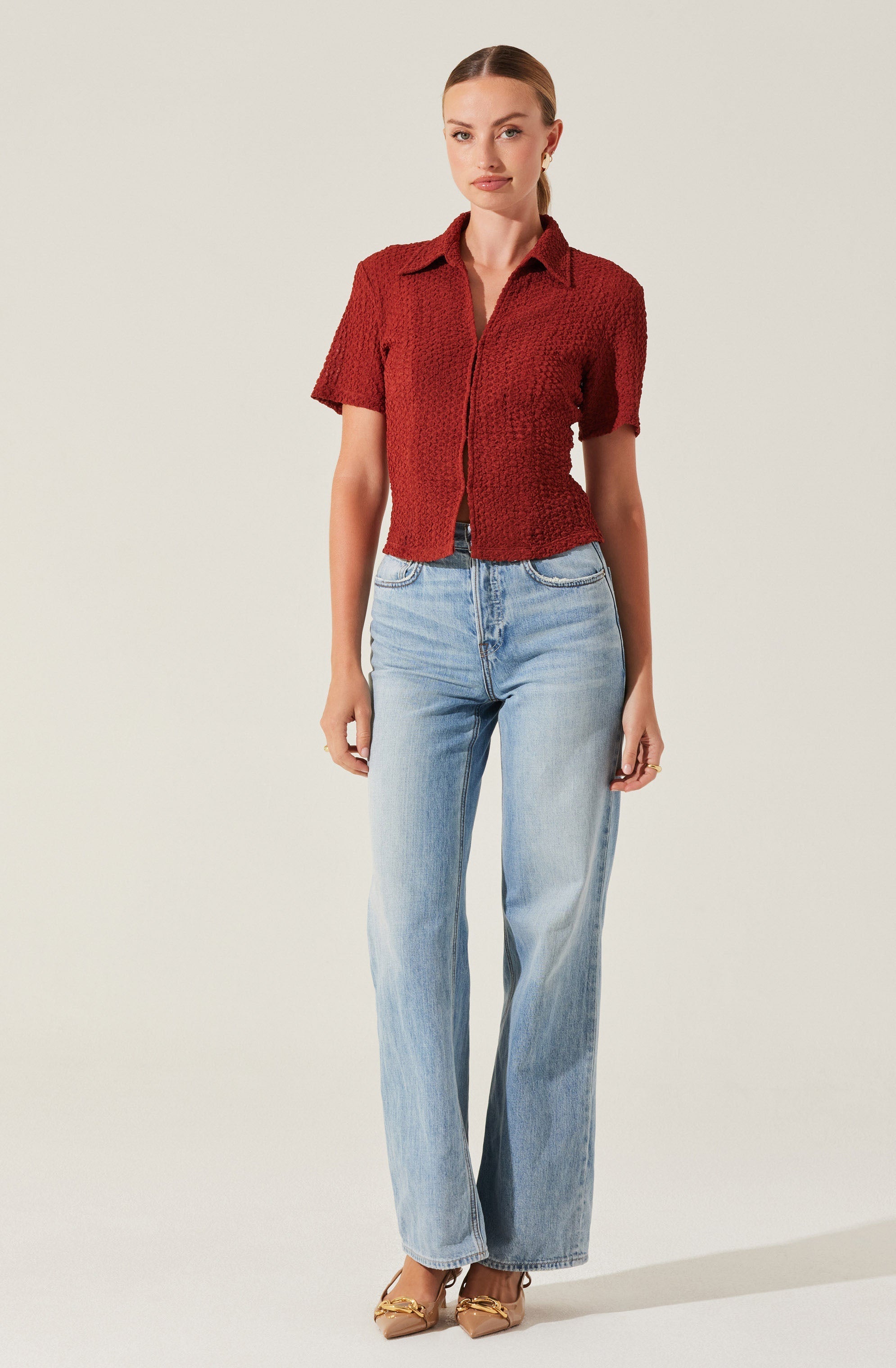 A woman is wearing a short-sleeved, textured red shirt with a collar and buttons down the front. She pairs it with light-wash, high-waisted, wide-leg jeans. She completes the outfit with beige pointed-toe heels with gold embellishments.