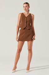 A woman is wearing a sleeveless brown top with a V-neckline and a row of buttons down the front, paired with a matching brown skirt that is above knee length. She is also wearing white high-heeled shoes and statement earrings.