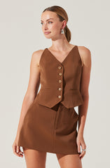 A woman is wearing a sleeveless, V-neck brown vest with a row of large, round beige buttons down the front. She pairs it with a matching brown skirt that has a front pleat. She accessorizes with large, white drop earrings and wears a silver ring on her right hand.
