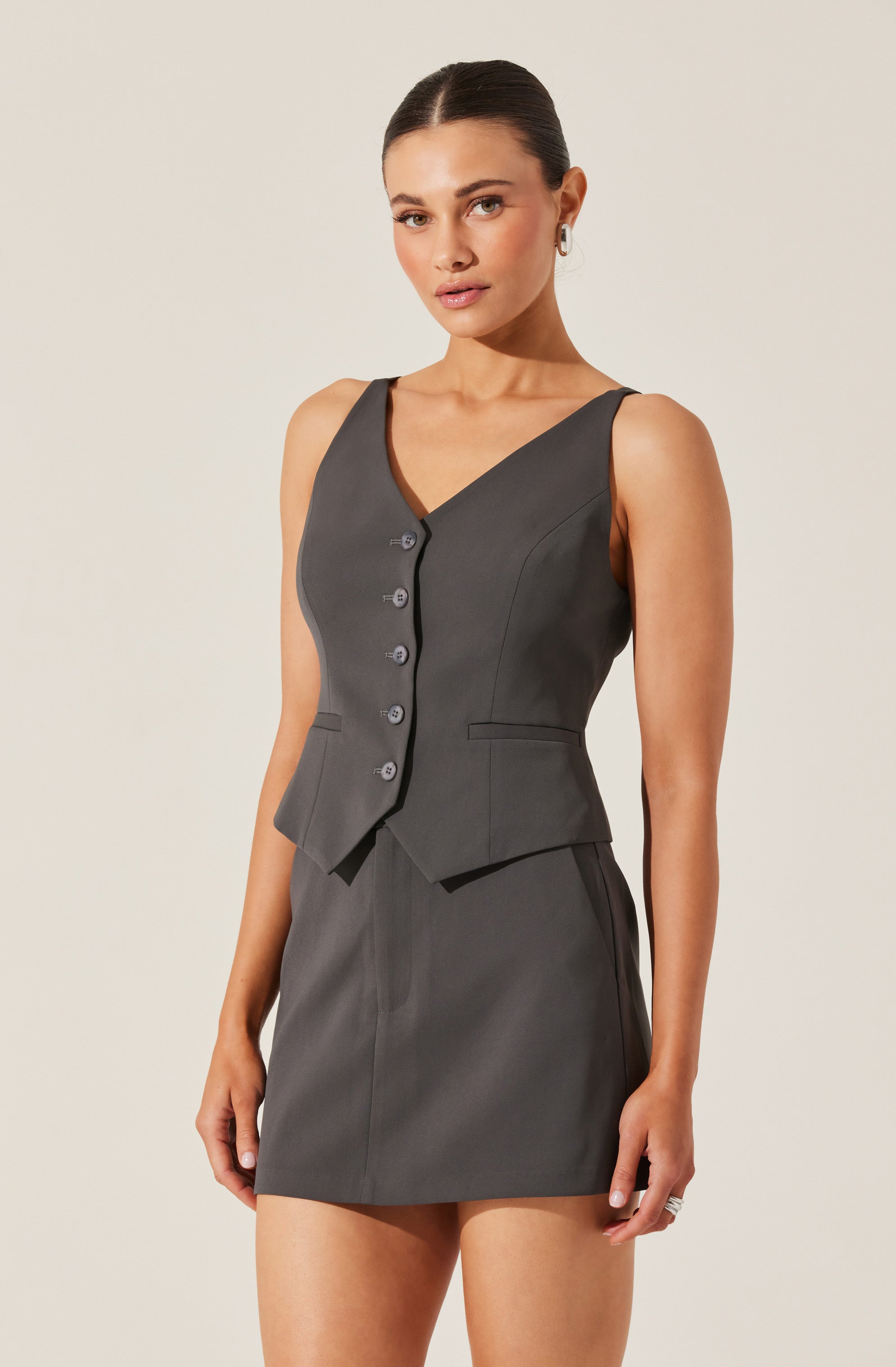 A woman is wearing a sleeveless, dark gray vest with a V-neckline and front buttons. The vest is tailored with princess seams and has a peplum-style hem. She pairs it with a matching dark gray high-waisted skirt that is fitted and falls to mid-thigh.