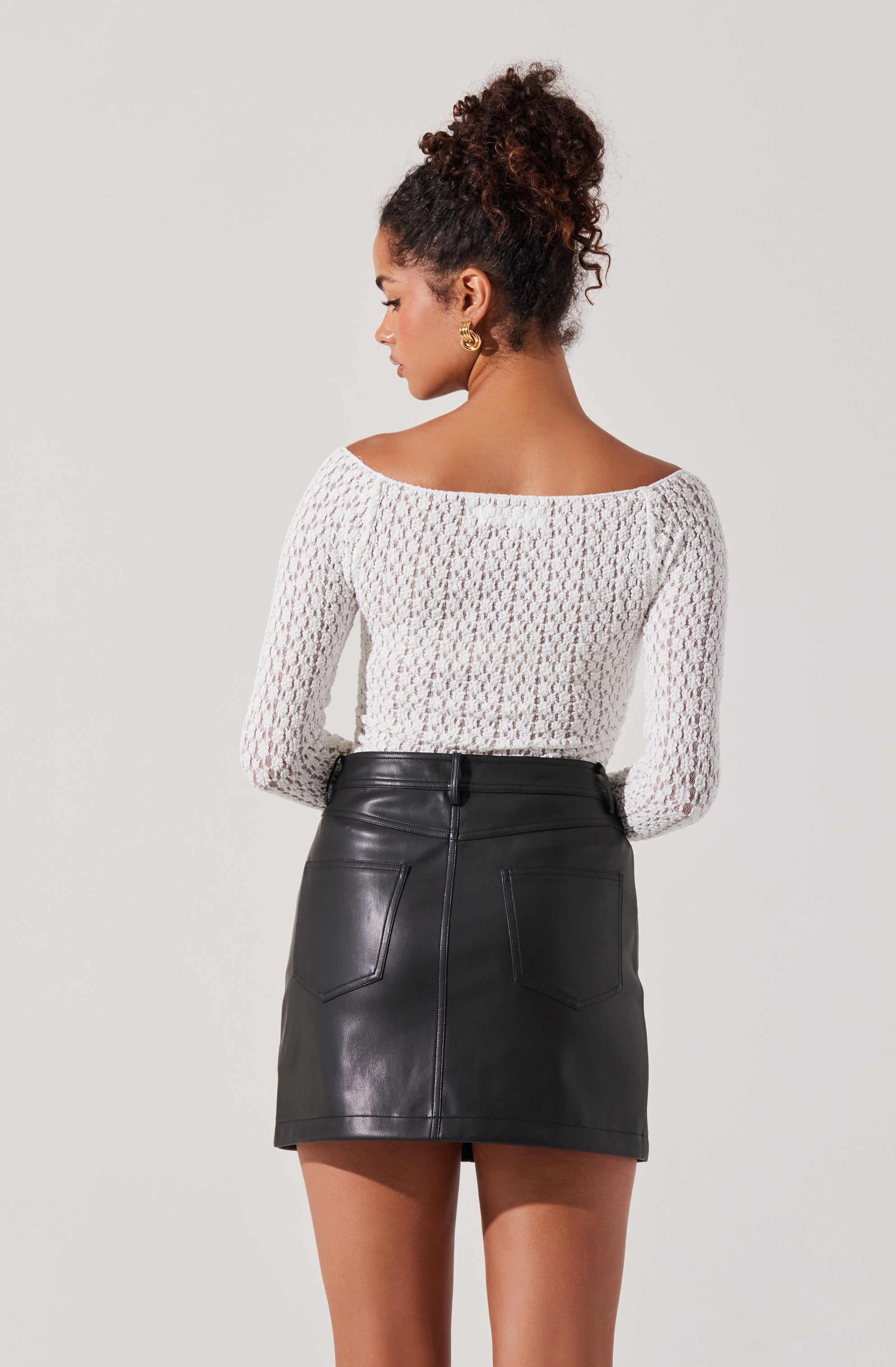 A woman is wearing a long-sleeve, white, open-knit top with a wide neckline that reveals her shoulders. She pairs it with a high-waisted, black leather mini skirt that features visible stitching and two back pockets.