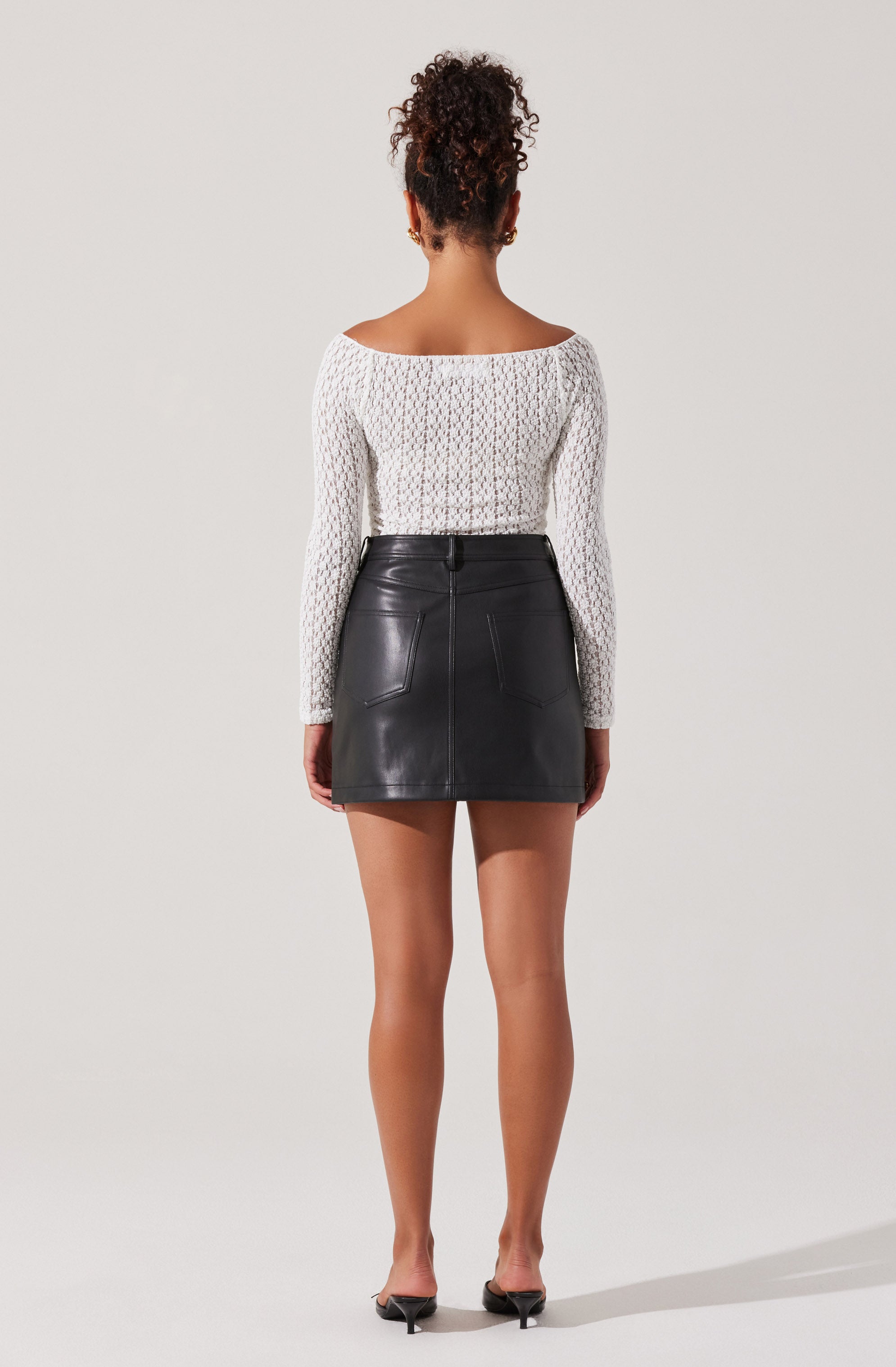 A woman is wearing a white, long-sleeved, textured knit top with a wide, rounded neckline, paired with a high-waisted black leather mini skirt that has visible stitching and front pockets. She is also wearing black open-toe heels.