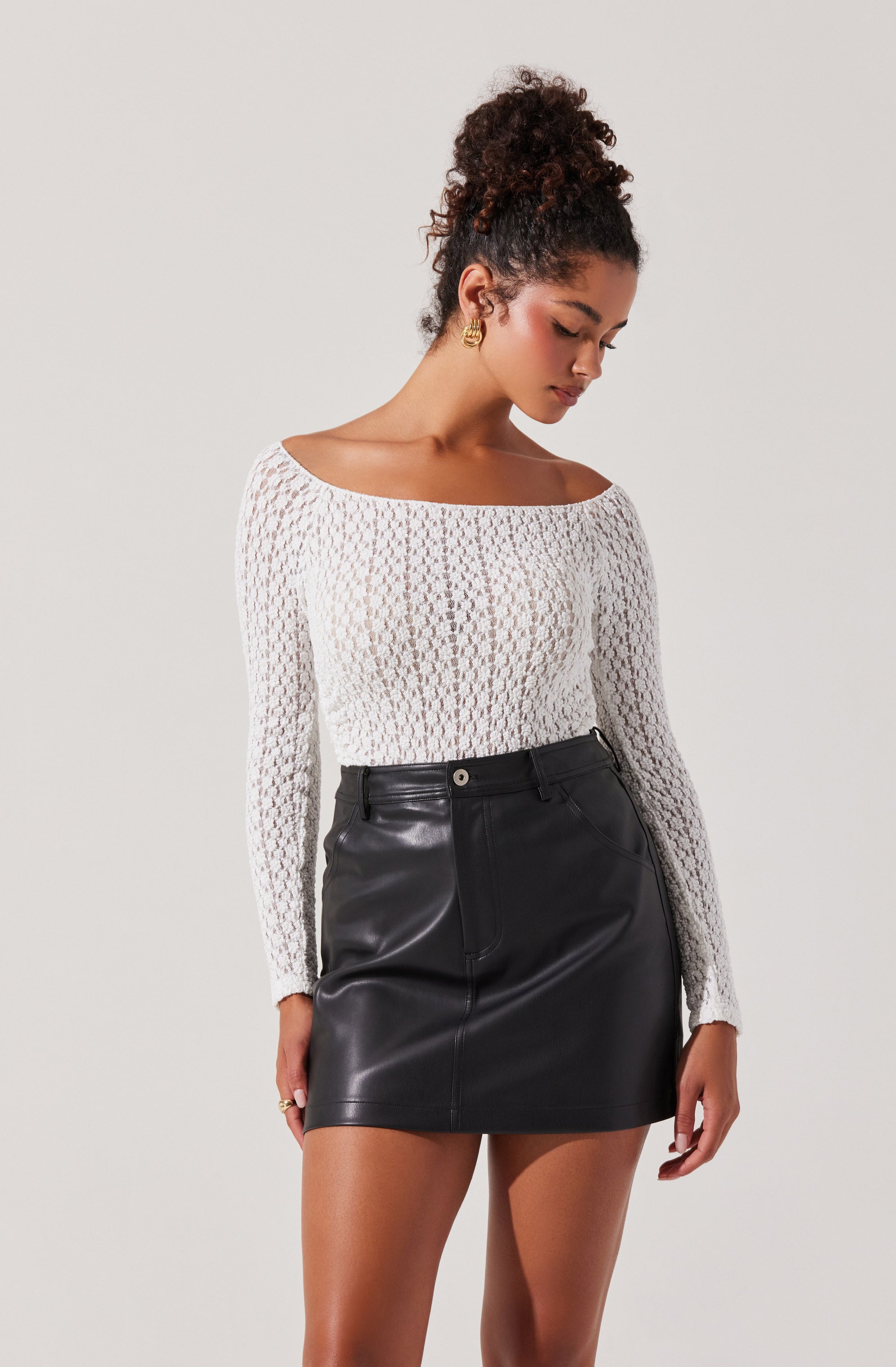 A woman is wearing a long-sleeved, white, open-knit sweater with a wide neckline and a black leather mini skirt. She has gold earrings and her hair is styled in a curly updo.