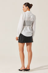 A woman is wearing a white long-sleeve button-up shirt with puffed sleeves and a collar. Over the shirt, she has on a light gray waist cincher or belt. She is also dressed in a black mini skirt made of a leather-like material and is wearing black open-toe high heels.
