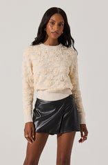 A woman is wearing an off-white, textured sweater with a raised, pom-pom-like pattern throughout. The sweater has a crew neckline and ribbed cuffs. She pairs it with a black faux leather mini skirt that has a slight A-line shape and a subtle wrap style.
