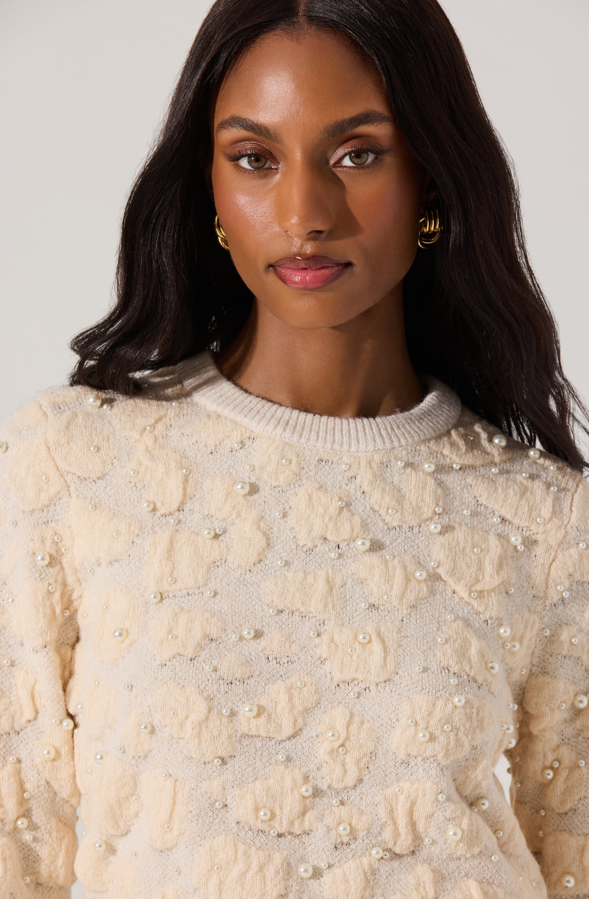 A woman is wearing a cream-colored sweater with textured, ruffled fabric and pearl embellishments scattered throughout. The sweater has a round neckline and long sleeves.