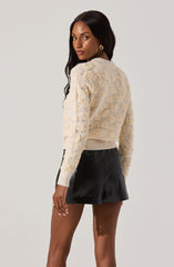 A woman is wearing a light-colored, textured sweater with decorative details like pearls or beads, along with a fitted waistband. She pairs it with a black leather mini skirt.