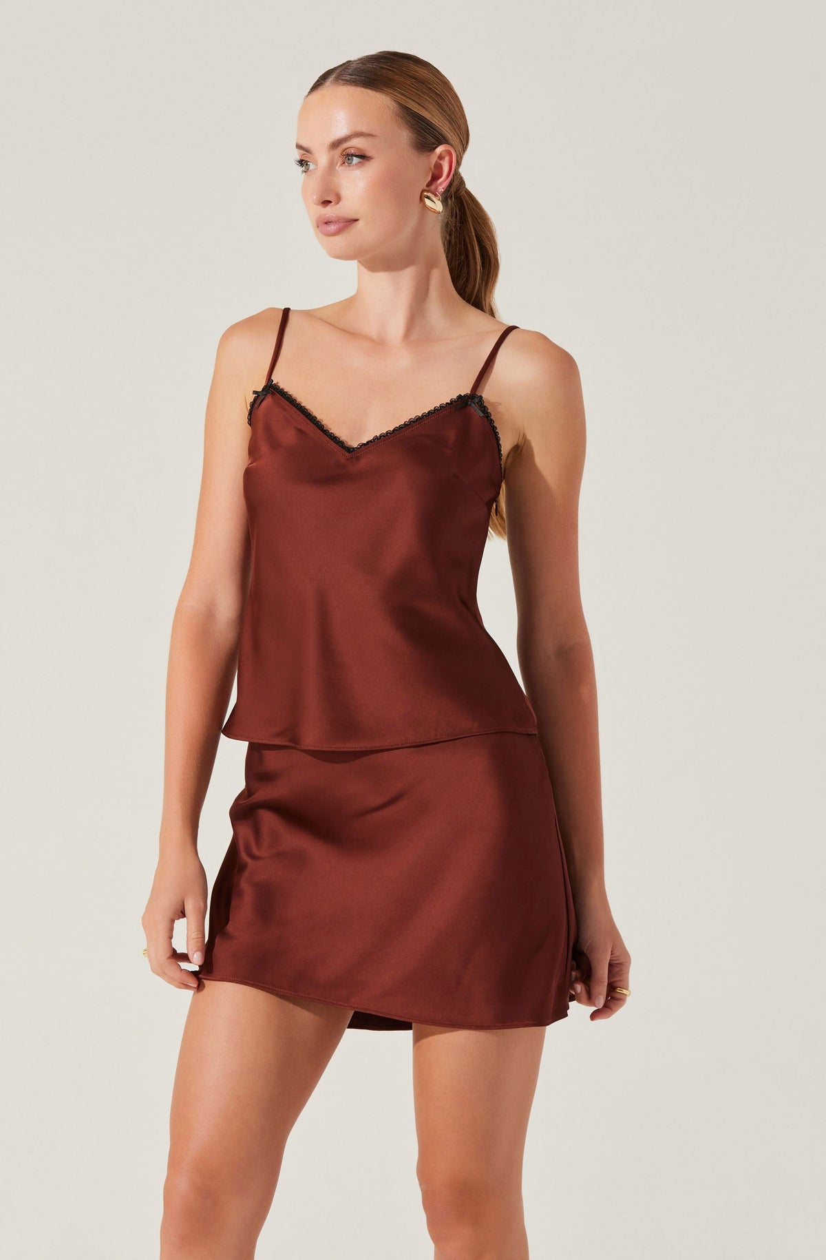 A woman is wearing a sleeveless satin camisole top with thin spaghetti straps and a V neckline accented with black lace trim. She pairs it with a matching satin mini skirt. The outfit is in a rich, reddish-brown color.