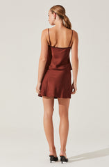 A woman is wearing a satin camisole and matching satin mini skirt. The camisole is a sleek, shiny fabric in a warm, reddish-brown color with thin spaghetti straps and a slightly loose fit. The skirt is also made of shiny satin material, fitted around the waist and flaring slightly toward the hem. She is wearing black high-heeled shoes.