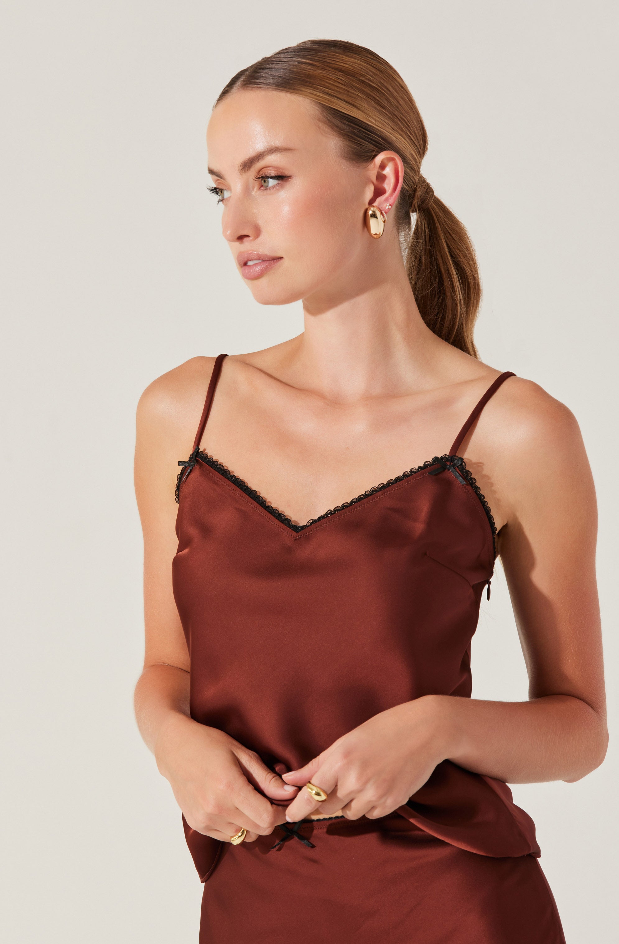 A woman is wearing a silky, satin-like camisole top in a deep burgundy color with thin spaghetti straps. The top features black lace trim along the V-neckline. She is accessorized with gold hoop earrings and multiple gold rings.