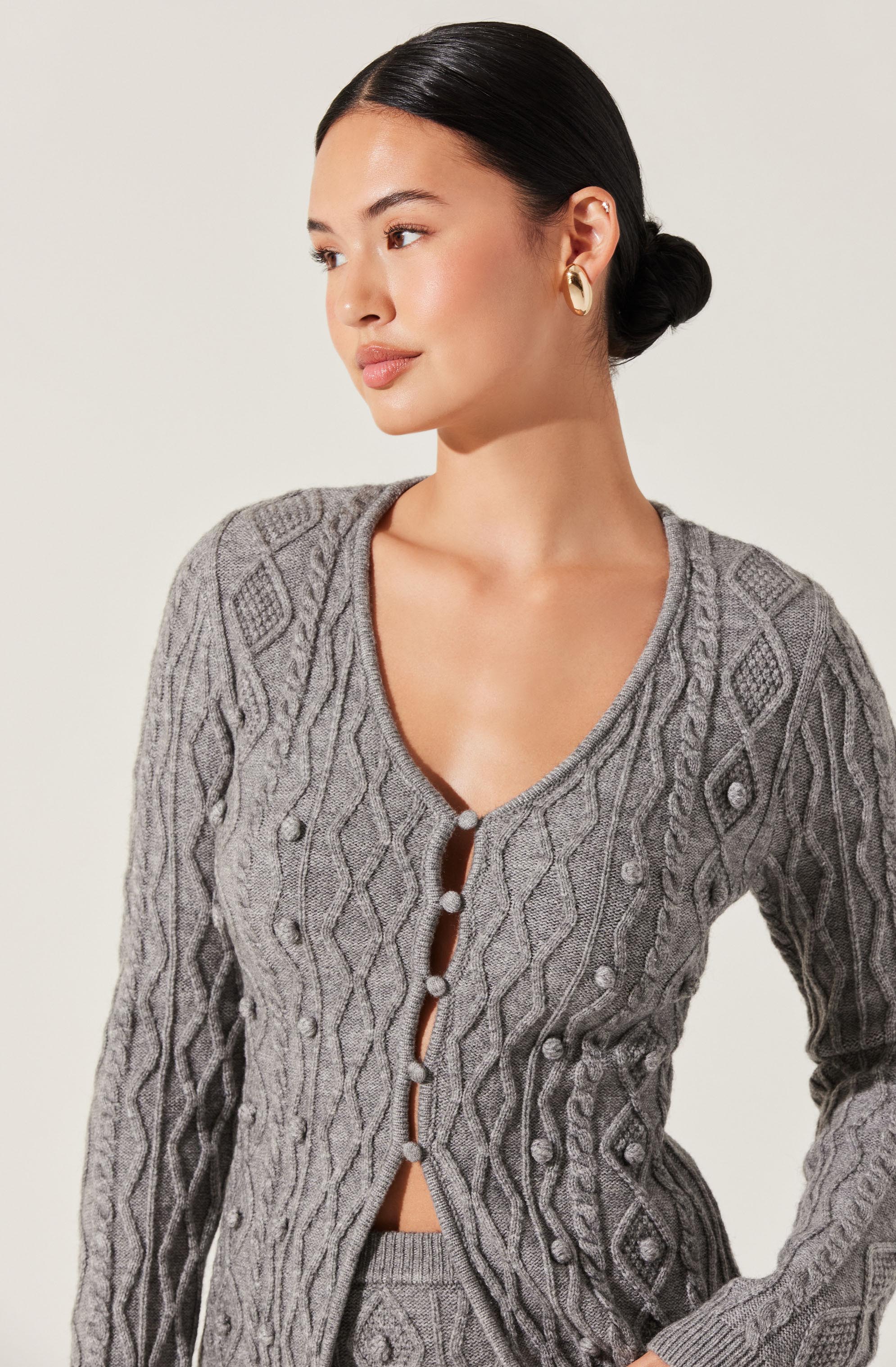 A woman is wearing a gray, long-sleeved, cable-knit cardigan with intricate diamond and braid patterns. The cardigan has a V-neckline with decorative buttons running down the front. She is also wearing gold hoop earrings.