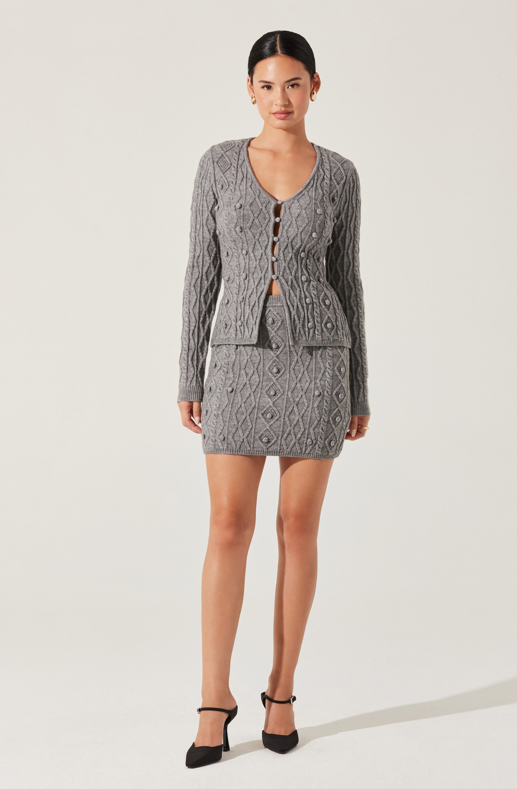 A woman is wearing a matching two-piece outfit consisting of a gray cable-knit cardigan with diamond patterns and a matching gray mini skirt. The cardigan has long sleeves, a round neckline with a button-up front featuring brown buttons, and is slightly cropped. The skirt is fitted and short, ending above the knees.