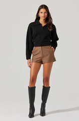 A woman is wearing a black long-sleeve, collared sweater paired with high-waisted brown shorts. She has on tall black boots that reach just below her knees.