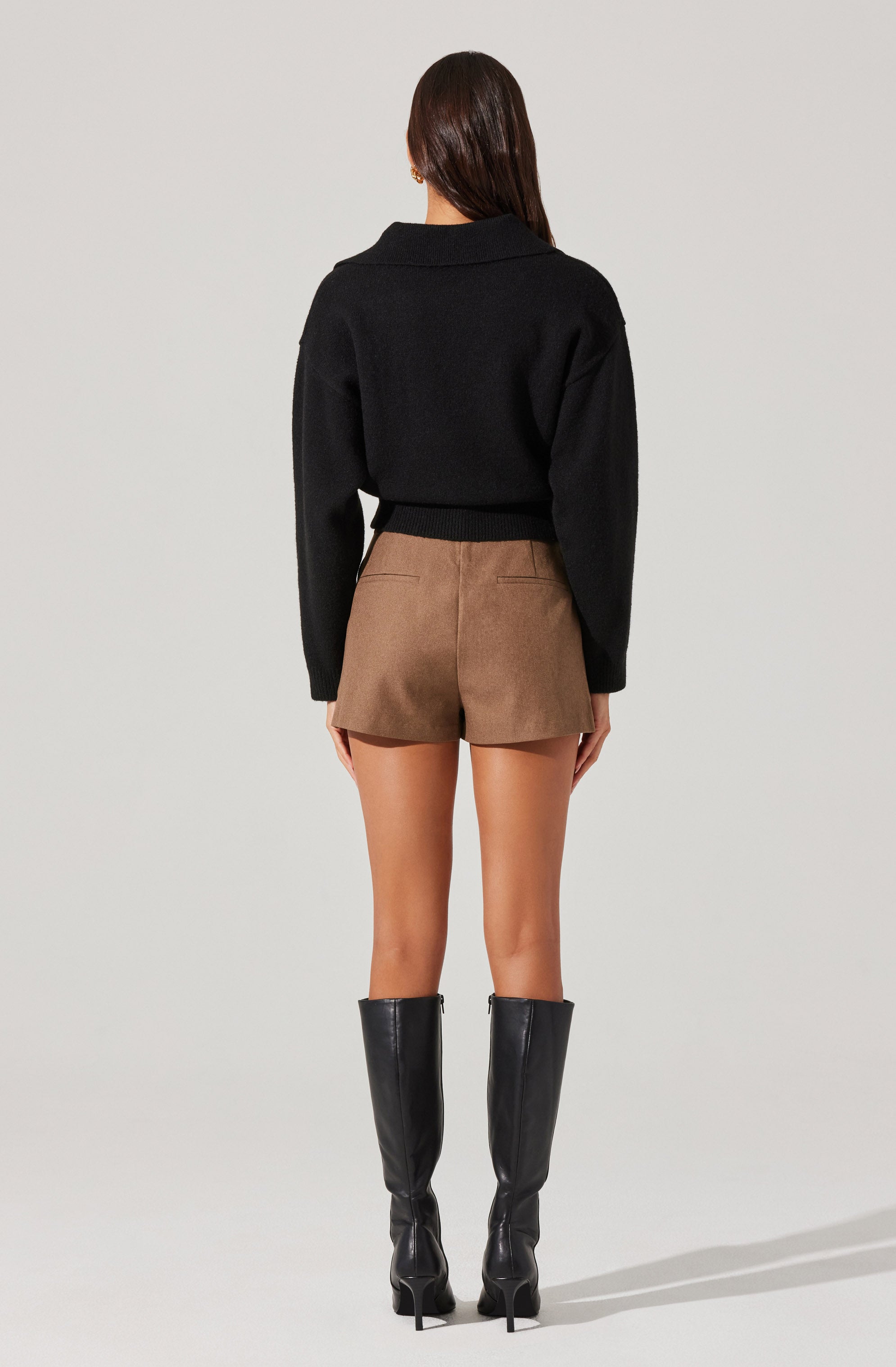 A woman is wearing a black, long-sleeved, cropped sweater with a wide collar, paired with high-waisted, brown shorts. She is also wearing tall, black knee-high boots with high heels.