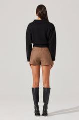 A woman is wearing a black, long-sleeved, cropped sweater with a wide collar, paired with high-waisted, brown shorts. She is also wearing tall, black knee-high boots with high heels.