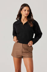 A woman is wearing a black long-sleeve sweater with a collar and a half-zip neckline. She pairs it with high-waisted, tailored brown shorts that have a front seam detail.