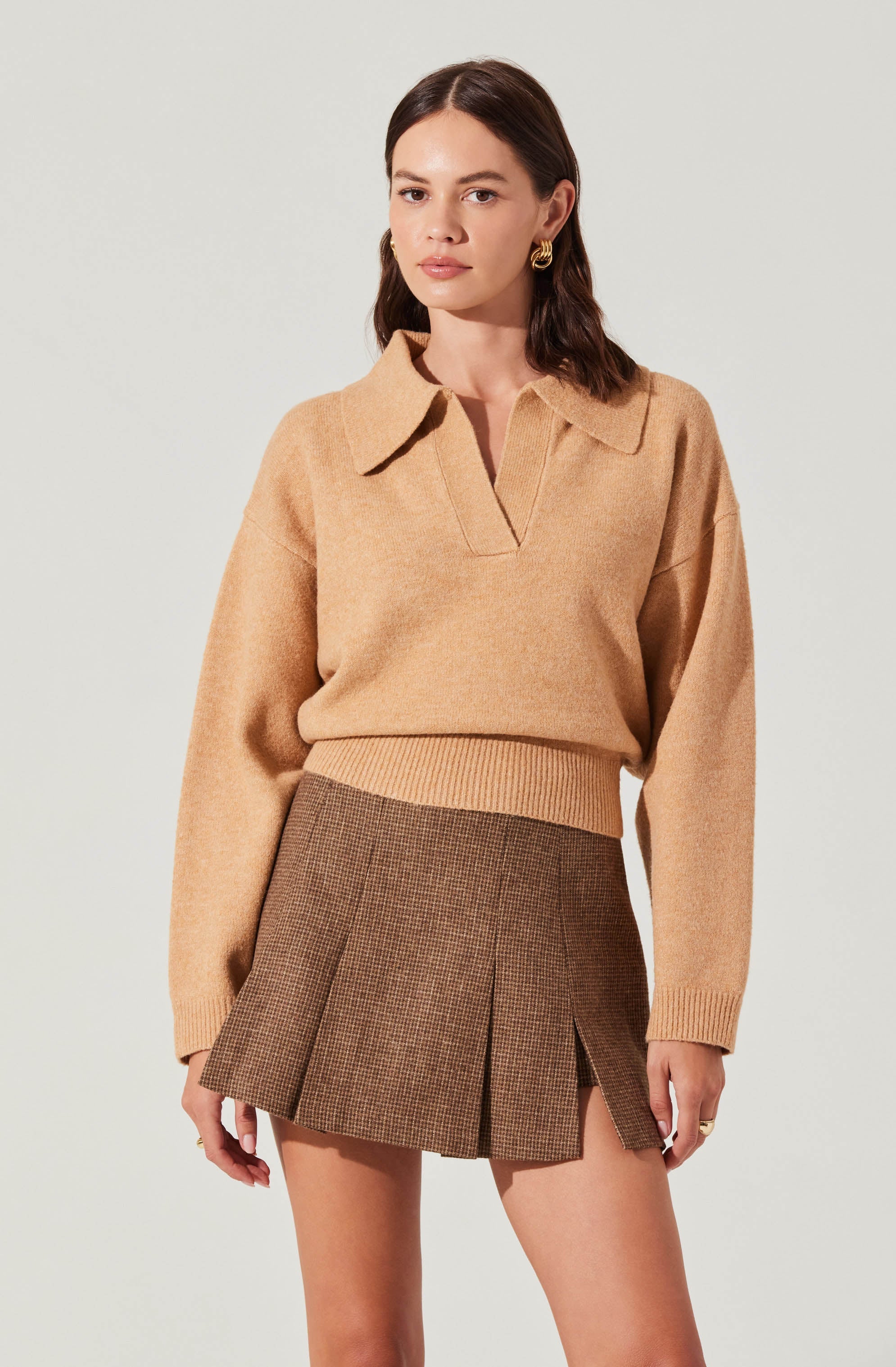 A woman is wearing a beige, long-sleeved sweater with a collar and a V-shaped neckline, paired with a high-waisted, brown plaid skirt featuring front pleats and a side slit.