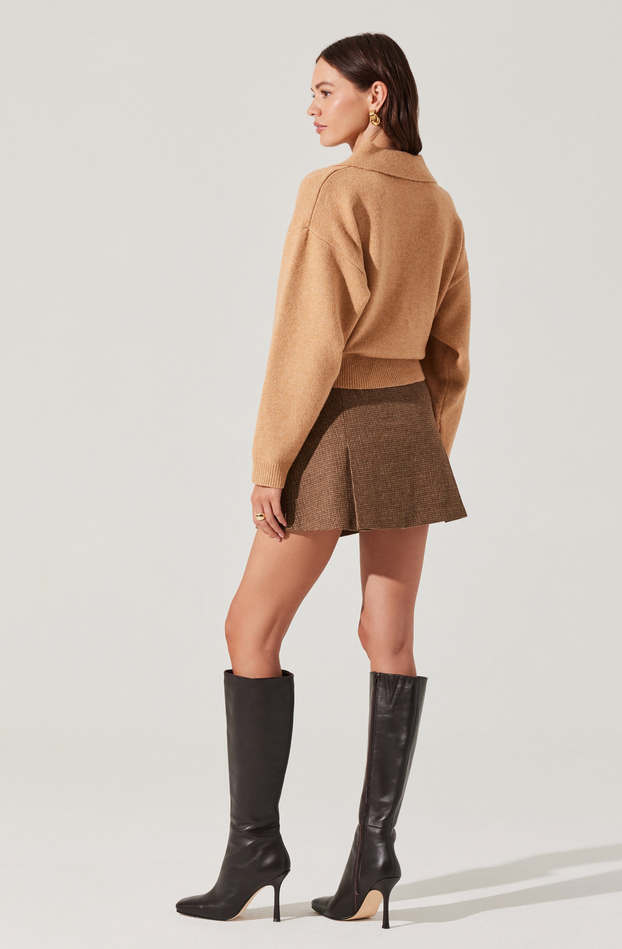A woman is wearing a camel-colored, oversized sweater with a collar and long sleeves. She pairs it with a short brown plaid skirt that features a front slit. She completes the outfit with tall black leather high-heeled boots.
