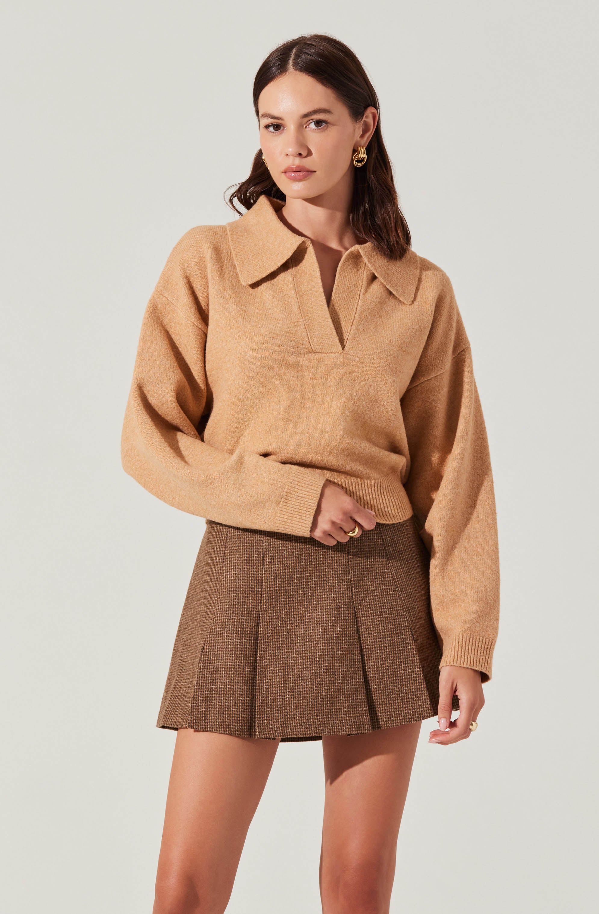 A woman is wearing a camel-colored, oversized sweater with a collar and a v-shaped neckline. She pairs it with a brown, high-waisted, plaid mini skirt that has pleats. She accessorizes with gold hoop earrings and gold rings.