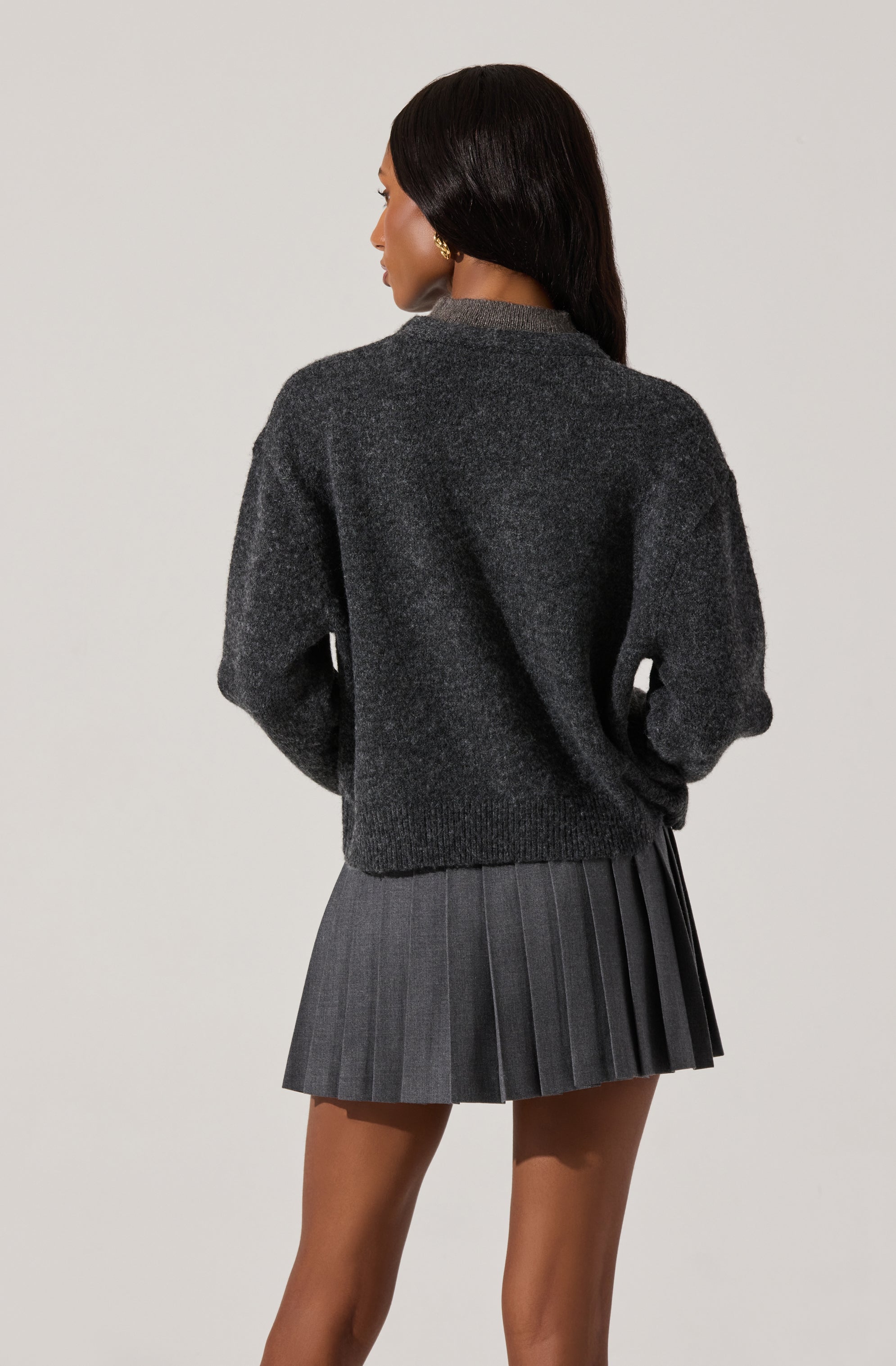 A woman is wearing a dark gray, fuzzy knit sweater with a relaxed fit. The sweater has a round neckline and long sleeves that are slightly puffed at the cuffs. She pairs it with a gray pleated mini skirt.