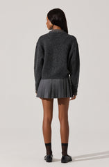 A woman is wearing a dark gray, long-sleeved, crewneck sweater that appears soft and cozy. She is also wearing a dark gray pleated skirt that falls above her knees, with evenly spaced pleats. Her outfit is completed with black ankle socks and black shoes.