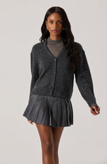 A woman is wearing a dark gray, button-up cardigan sweater over a matching gray knit top. She has on a pleated gray skirt with vertical stripes, and she is accessorized with gold hoop earrings.