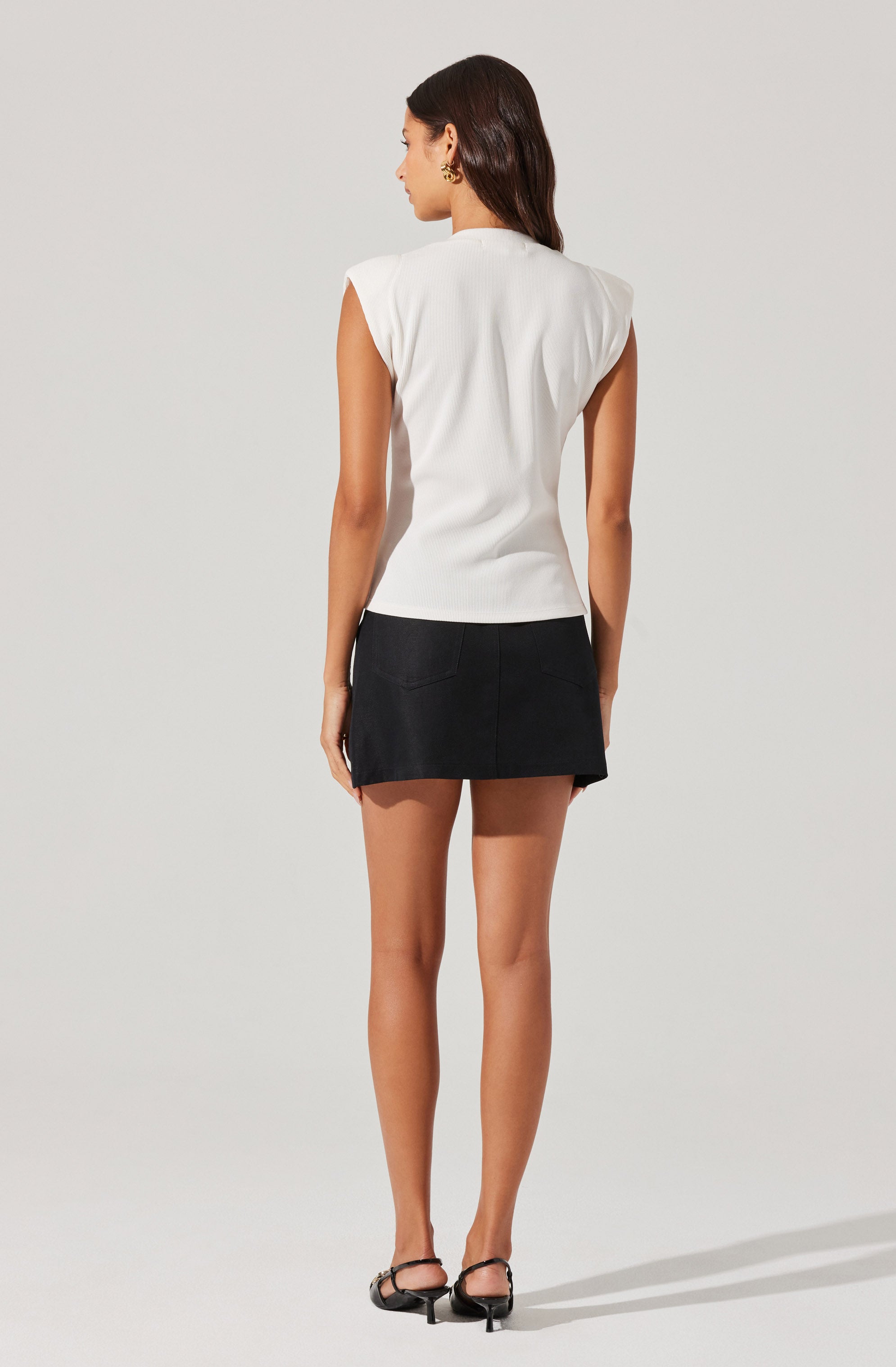 A woman is wearing a sleeveless white top with padded shoulders and a black, above-the-knee skirt. She is also wearing black open-toe heeled sandals.