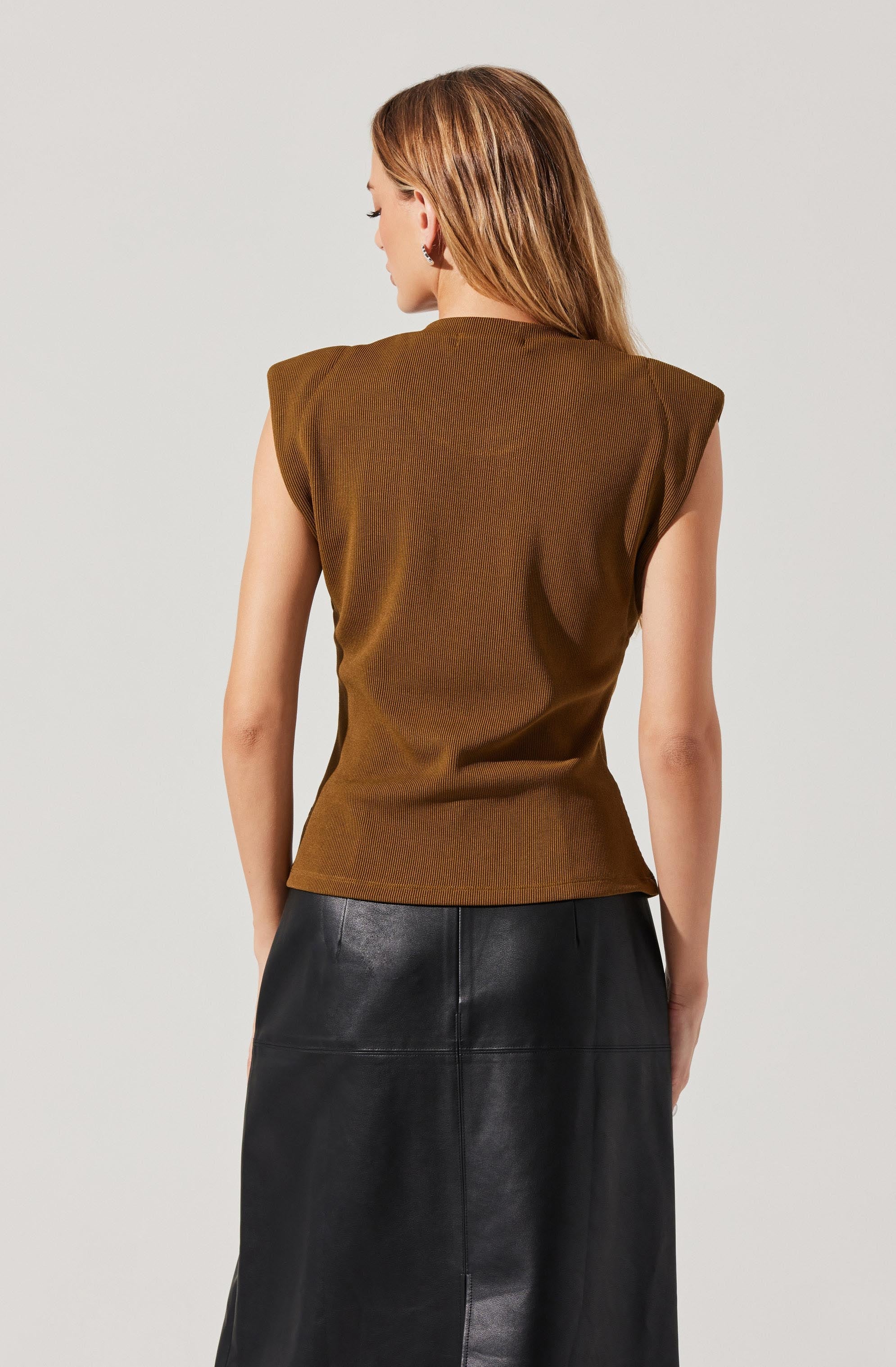 A woman is wearing a brown sleeveless top with padded shoulders and a high neckline. She pairs it with a long black leather skirt.