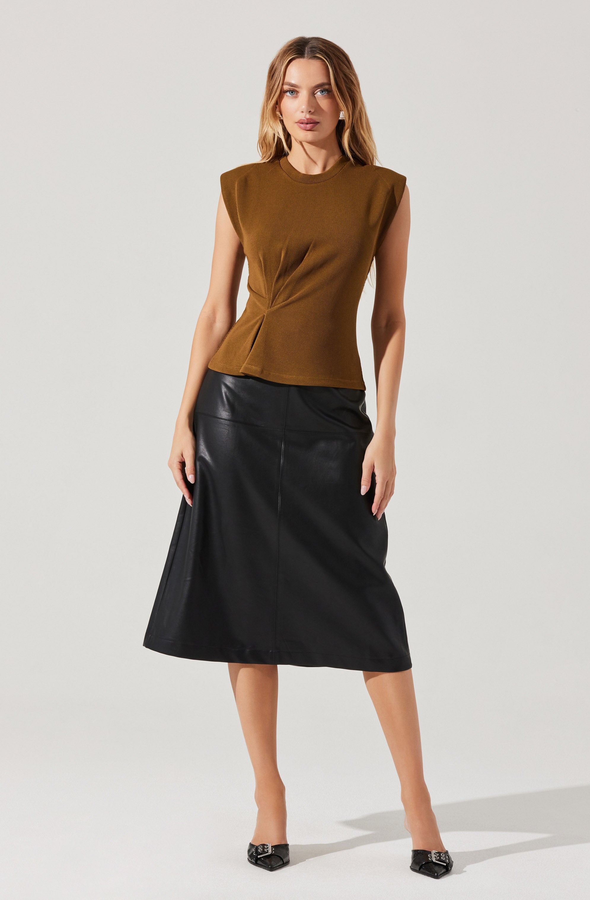 A woman is wearing a sleeveless, structured, brown top with exaggerated shoulder details and a subtle pleat at the waist. She pairs it with a black, midi-length, leather skirt that has a slight A-line shape. She completes the outfit with pointed-toe black shoes featuring decorative embellishments.