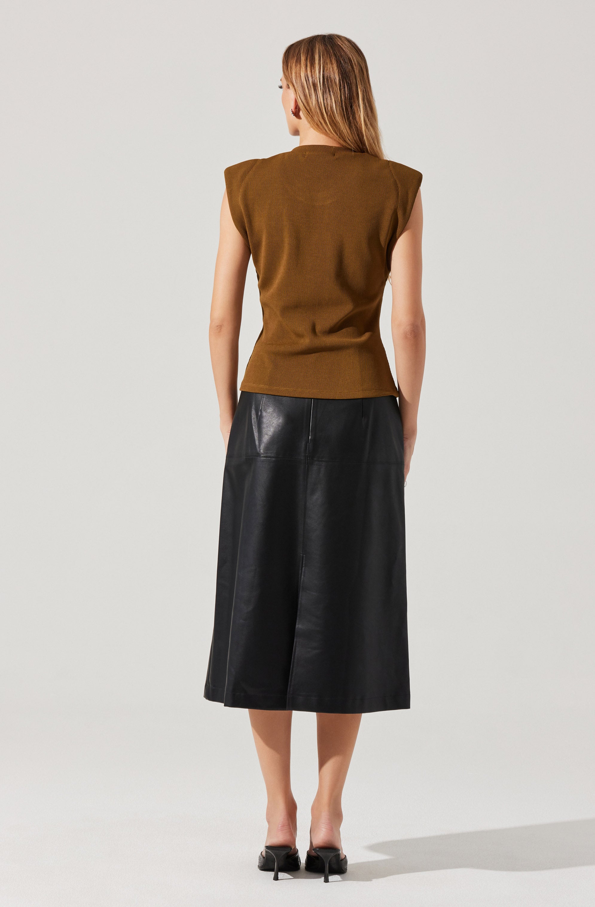 A woman is wearing a sleeveless brown top with padded shoulders and a high neckline. She pairs it with a black, high-waisted, midi-length leather skirt that has a slightly flared silhouette. She is also wearing black, open-toe, mid-heeled sandals.